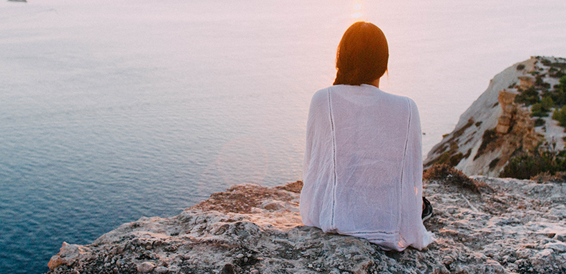 A woman is relaxing on a large rock while viewing the sea.