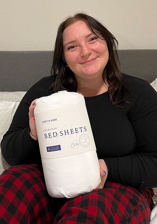 A smiling female customer is holding a packaged set of Cosy House Collectionâs Everyday Bed sheets.