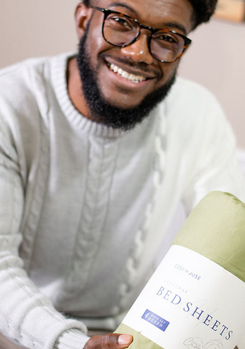 A smiling male customer is holding a packaged set of Cosy House Collectionâs Everyday Bed sheets.