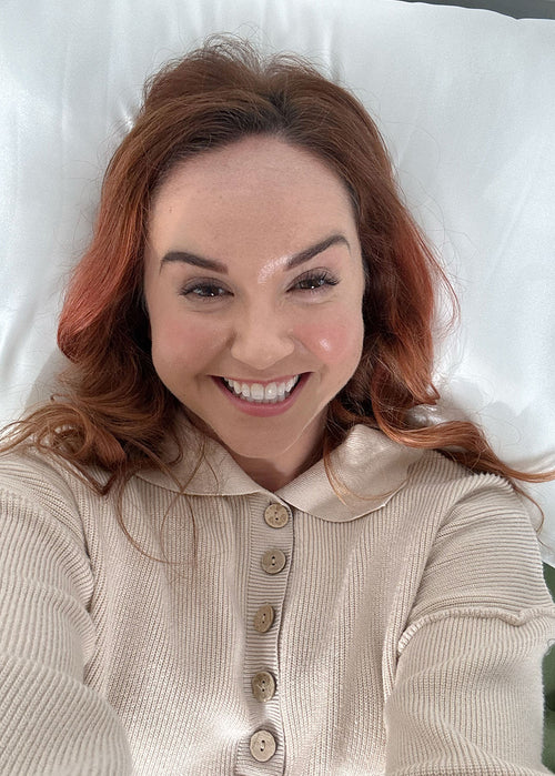 Woman with red hair lying on a white silk pillowcases, wearing a beige cardigan.