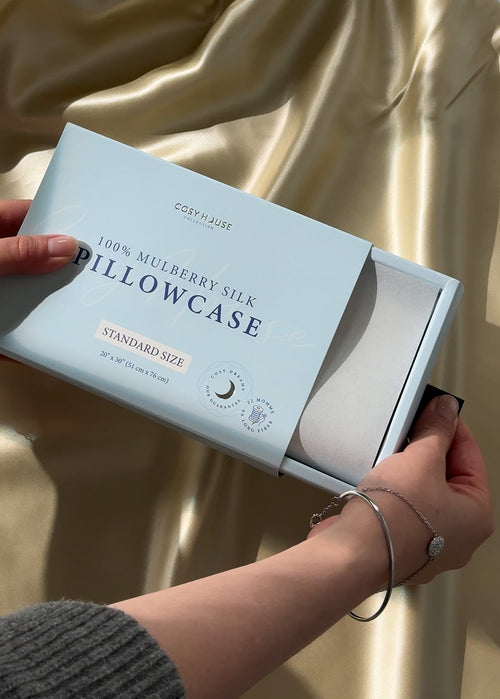Cosy House 100% Mulberry Silk Pillowcase packaging held by a person against a gold curtain background.