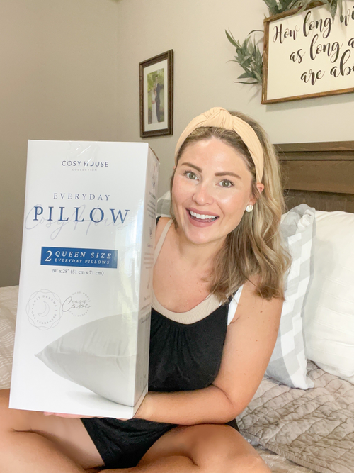 A satisfied customer sitting on the bed showing a packaged pair of Everyday Down Alternative Pillows.