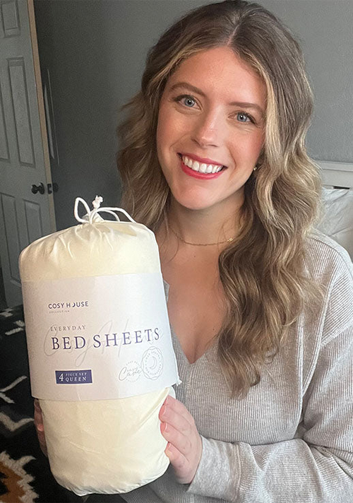 A happy female customer is holding a packaged set of Cosy House Collectionâs Everyday Bed sheets.