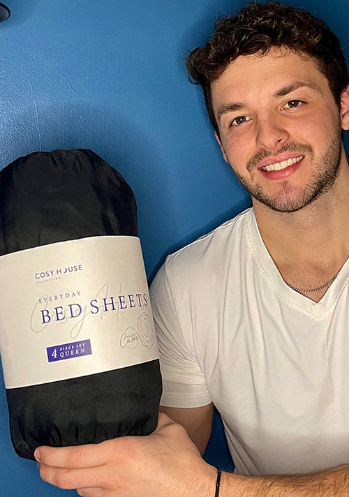 A happy male customer is holding a packaged set of Cosy House Collectionâs Everyday Bed sheets.