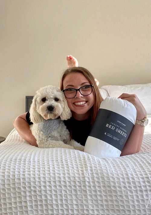 A smiling customer cuddling her dog and a packaged set of Cosy House Collectionâs Luxury bed sheets on a cozy bed.