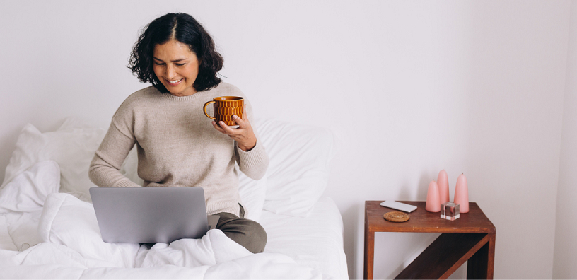 A woman is using her laptop with a cup of coffee on the bed.