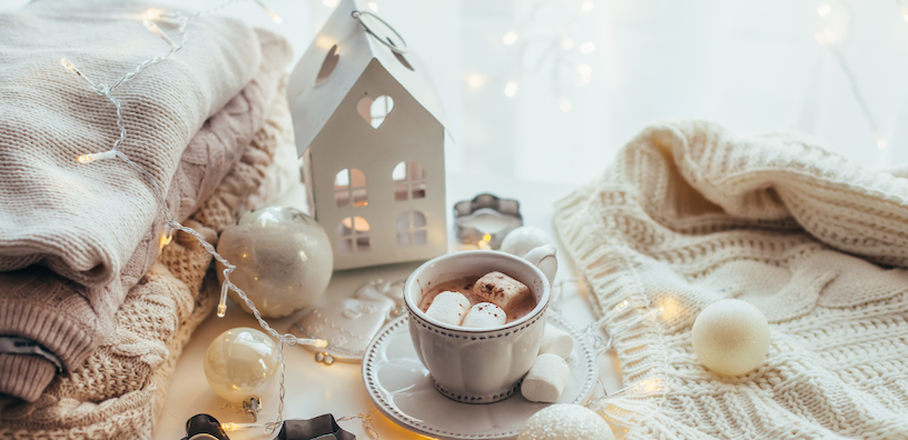 Cozy winter scene with hot chocolate, marshmallows, knitted sweaters, and decorative lights.