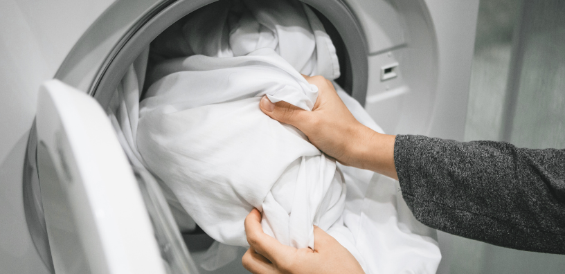 Two hands holding a bright, white bedding linen up to place it into a washing machine.