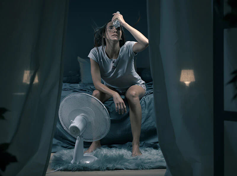 A woman sits on her bed, with a fan gently blowing air around her.