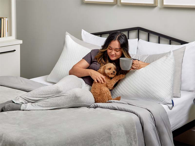 A woman relaxes on her bed, smiling as her dog cuddles beside her, creating a cozy and heartwarming scene.