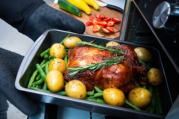 A person is using oven mitts to hold a pan filled with a golden roast chicken and crispy potatoes, ready to serve a delicious meal.