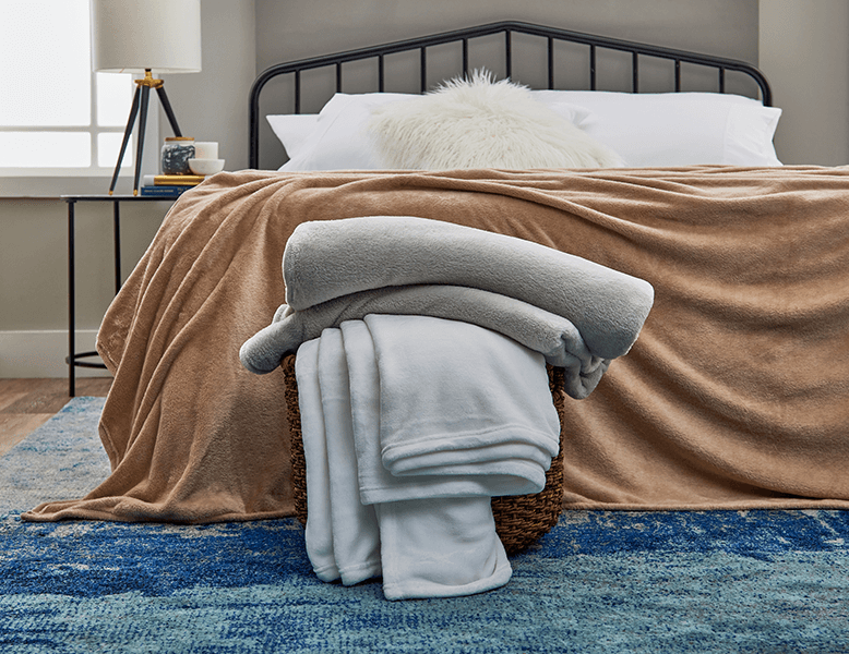 A comfortable bed adorned with a colorful fleece blanket. A basket containing other fleece blankets is also in front of the bed.