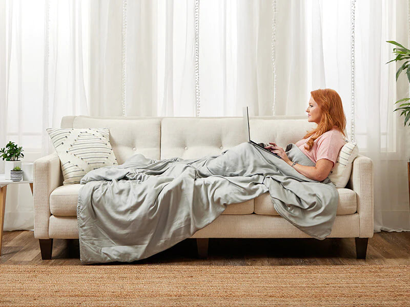 A woman with a blanket is seated on a couch, engaged with her laptop, embodying a casual and cozy home workspace vibe.
