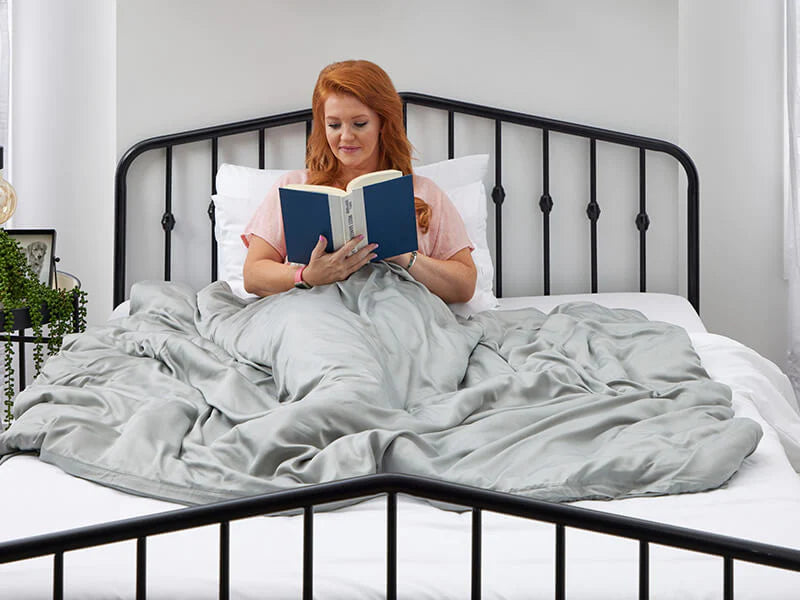A woman relaxing on her bed, deeply focused on a book, with a comfy blanket draped over her for warmth and comfort.