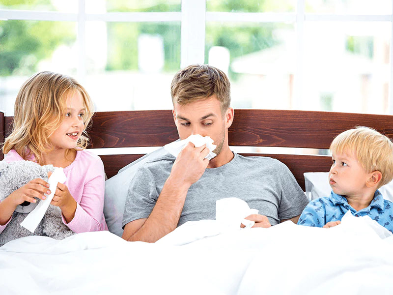 A man and two kids are snuggled in bed with tissues scattered around.